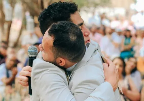 Dos hombres se abrazan en una boda, uno de ellos lleva un micrófono en la mano. Plantilla de discurso de boda, La Discursería.